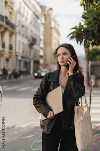 young freelancer in black leather jacket holding laptop and talking on smartphone on street in paris.