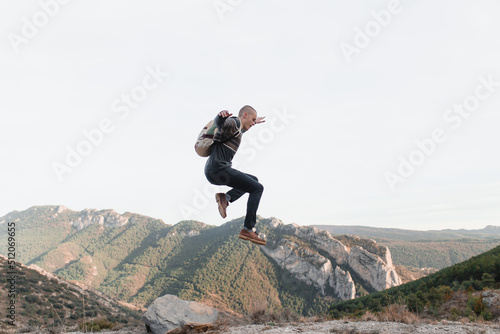 Shaved guy with backpack jumps with mountains in the background