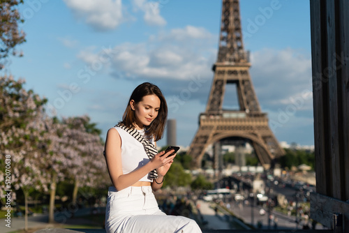 Photography young woman in stylish outfit using smartphone while sitting near eiffel tower in paris