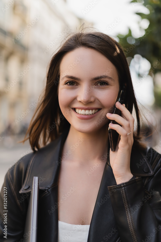 portrait of cheerful woman in black leather jacket holding laptop and talking on smartphone on street in paris. portrait of cheerful woman in black leather jacket holding laptop and talking on smartphone on street in paris.