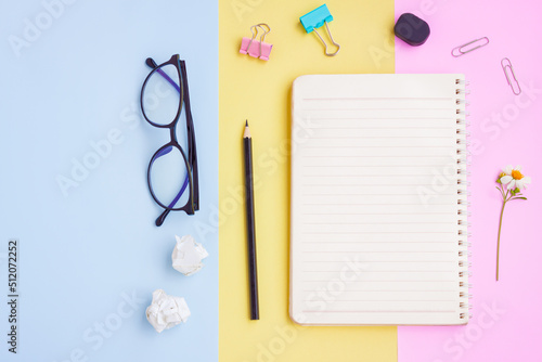 Notebook ,Eyeglasses,Black pencil,Eraser,Paper Clips and White Flower on Pink,Yellow and Blue Background