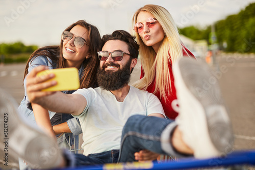 Obraz na płótnie Cheerful ethnic guy sitting in pushcart and taking selfie with female friends