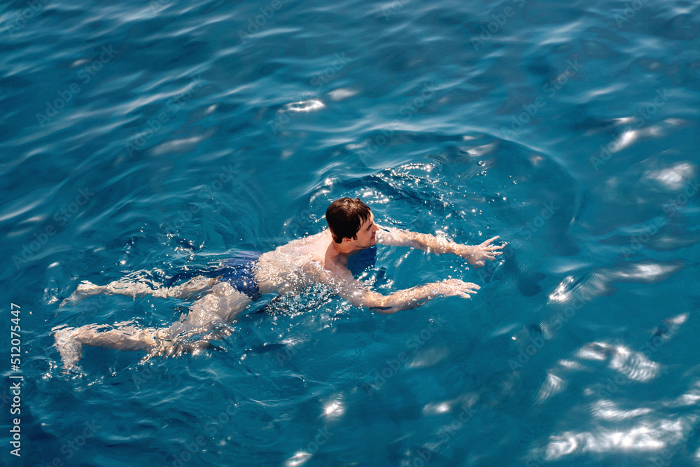 The guy swims in the blue calm sea water or ocean. Gentle sun glare on the surface. The concept of vacation, travel and enjoyment of a quiet life. Soft selective focus.