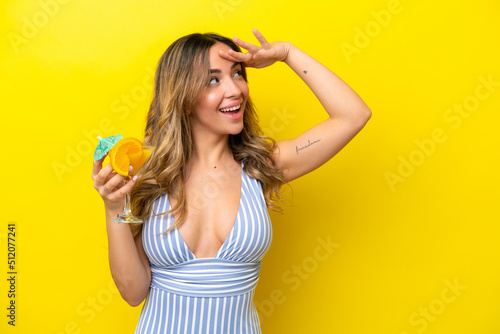 Young caucasian woman isolated on yellow background in swimsuit and holding a cocktail