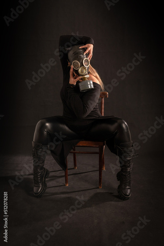 A girl in a full gas mask, latex pants, boots and robes is sitting on a chair on a black background in a pretentious pose with her head in her hands. Fetish, BDSM, perversion, bio o radiation hazard