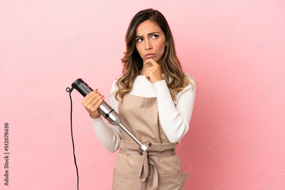 Young woman using hand blender over isolated pink background having doubts and thinking