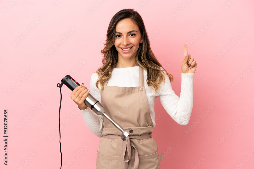Young woman using hand blender over isolated pink background pointing up a great idea Young woman using hand blender over isolated pink background pointing up a great idea