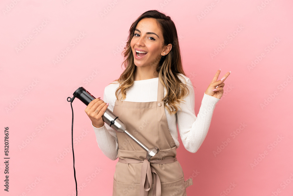 Young woman using hand blender over isolated pink background smiling and showing victory sign Young woman using hand blender over isolated pink background smiling and showing victory sign