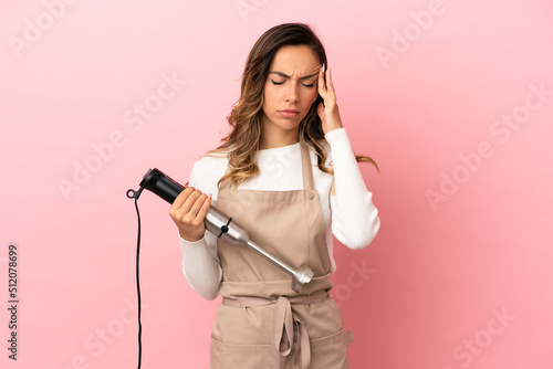 Young woman using hand blender over isolated pink background with headache