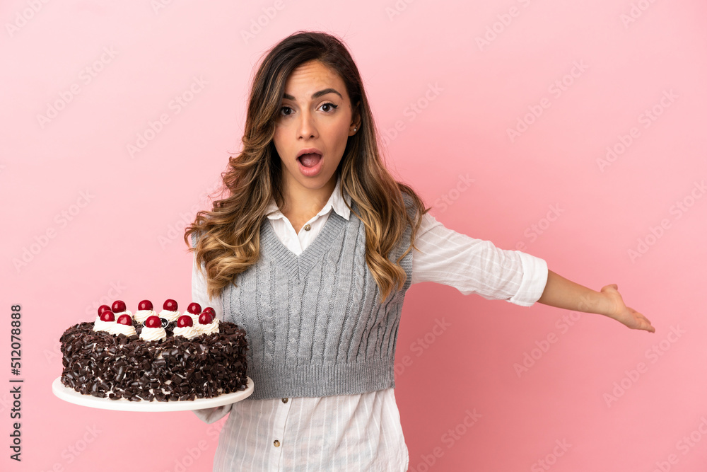Young woman holding birthday cake over isolated pink background extending hands to the side for inviting to come Young woman holding birthday cake over isolated pink background extending hands to the side for inviting to come