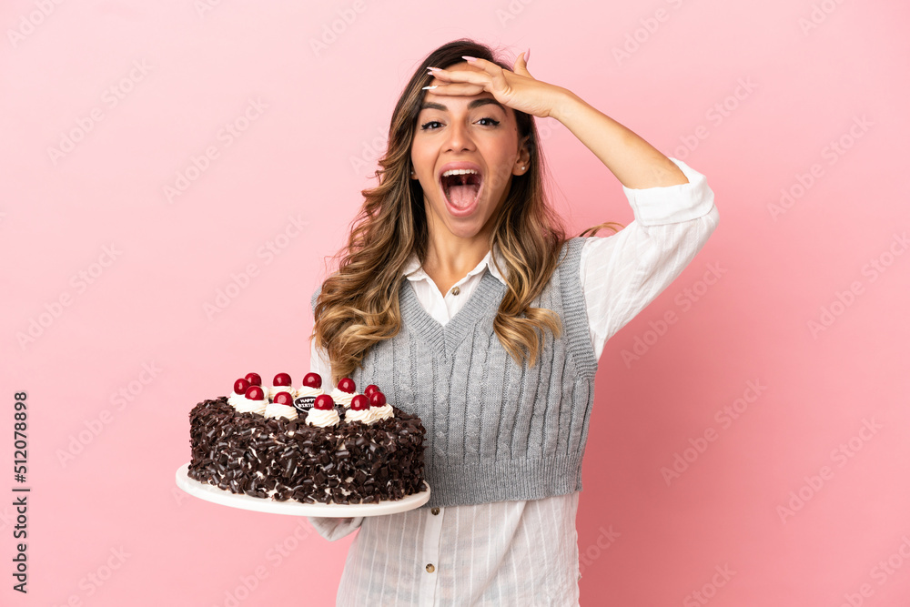 Young woman holding birthday cake over isolated pink background doing surprise gesture while looking to the side Young woman holding birthday cake over isolated pink background doing surprise gesture while looking to the side