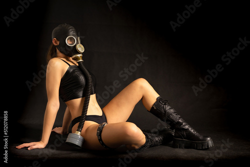 Seductive girl fetish model with a slim figure poses sitting in a black gas mask, black leather or latex sexy underwear and leather boots on a black background. BDSM, perversion, fetish, air pollution