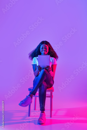 Attractive millennial black female working on tablet pc, chatting or video conferencing on web in neon light, copy space
