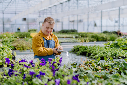 Happy young employee with Down syndrome working in garden centre, watering plants with a shower head and hose.