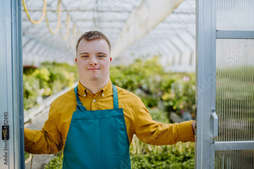 Young employee with Down syndrome working in garden centre, looking at camera and standing in door of greenhouse photo