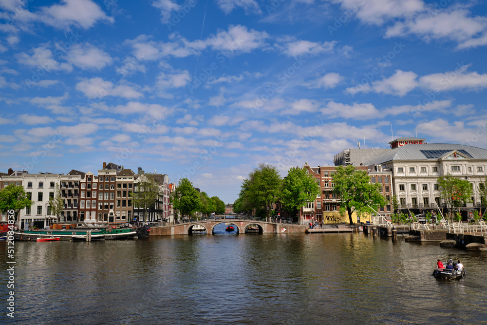 Amsterdam, Netherlands, 11 May 2022 - Panoramic.View along the Amstel river with bridge, Nieuwe Herengracht and the city skyline. Blue skies in spring sunshine.
