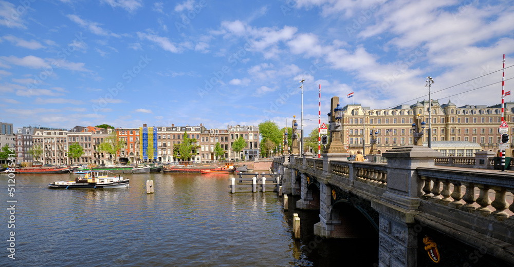 Naklejka premium Amsterdam, Netherlands, 11 May 2022 - Panoramic.View of bridge spanning the Amstel river with passenger boats and the city skyline. Blue skies in spring sunshine.