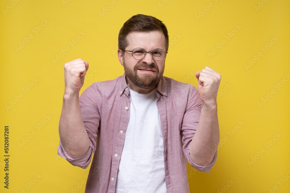 Man with glasses is upset by the losing, he makes a displeased grimace and holds his fists. Yellow background. Man with glasses is upset by the losing, he makes a displeased grimace and holds his fists. Yellow background.