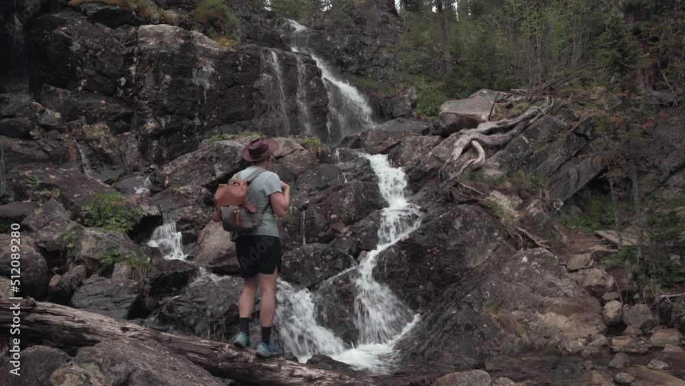 Tourist in hat with backpack stands with his back and looks at waterfall Tourist in hat with backpack stands with his back and looks at waterfall