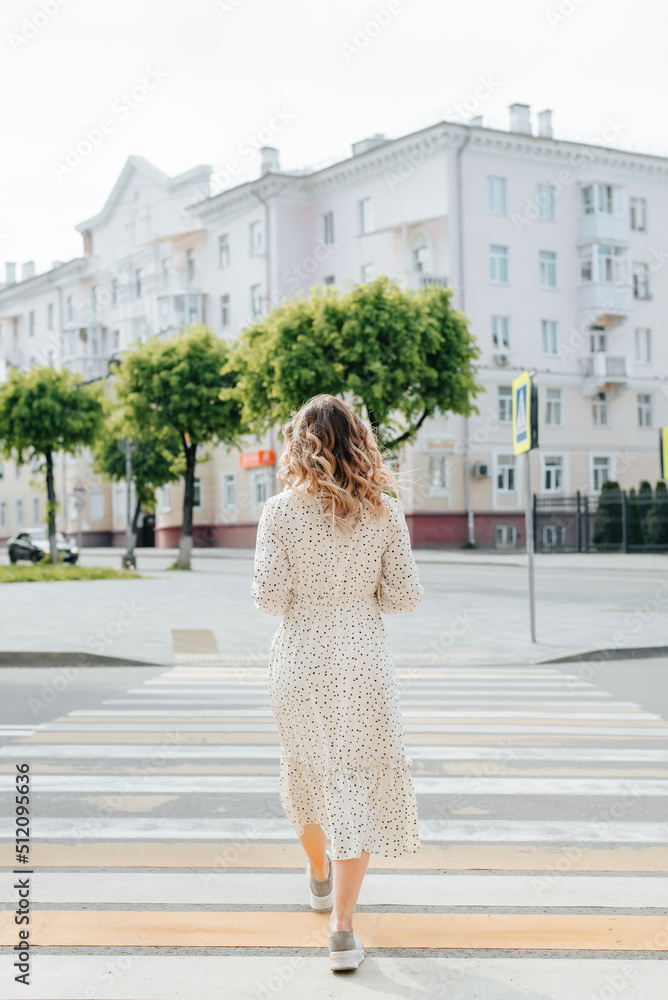 Stylish young woman with curly hairstyle crossing road outdoors, back view. Single slender woman wearing white summer dress walks along pedestrian crossing on street in city