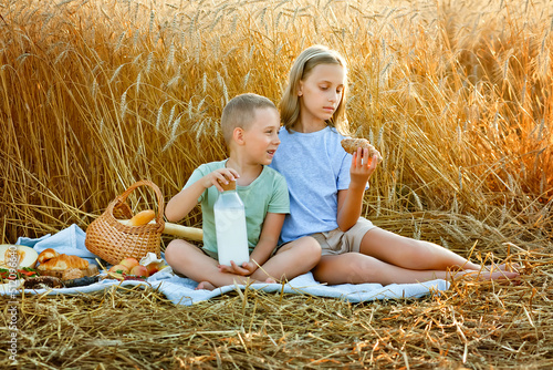 A boy and a girl are sitting on a blanket in a wheat field. The child eats in the fresh air. A walk and a picnic of friends in nature. They eat croissants and drink milk.