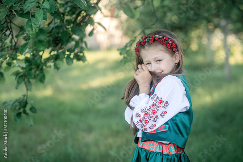little girl in a national ukrainian costume in an embroidered shirt and a red wreath  is touching her hair in the garden 