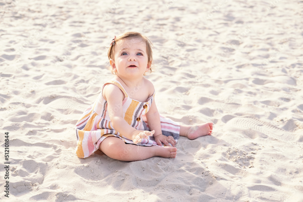 happy baby girl sitting on the beach on sand wearing striped summer dress on sunny day happy baby girl sitting on the beach on sand wearing striped summer dress on sunny day