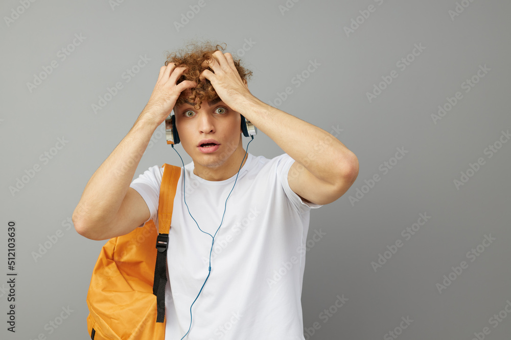 a puzzled student guy stands on a gray background in a white T-shirt and listening to music with headphones clutched his head looking at the camera with his eyes wide open a puzzled student guy stands on a gray background in a white T-shirt and listening to music with headphones clutched his head looking at the camera with his eyes wide open