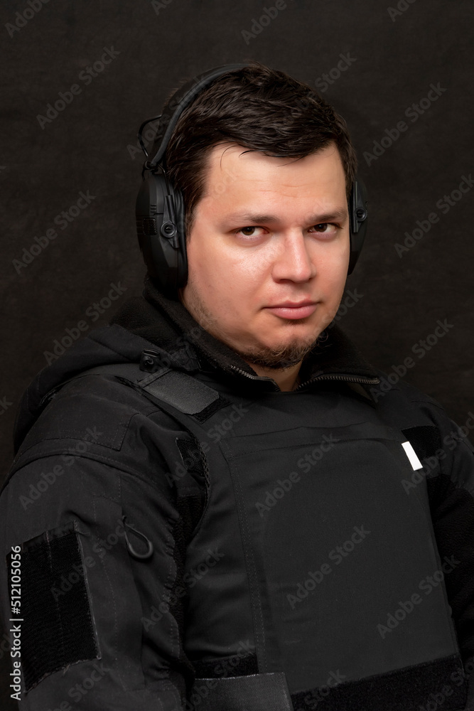 Obraz premium Portrait of a man 25-30 years old in a bulletproof vest and tactical headphones for shooting on a black background.