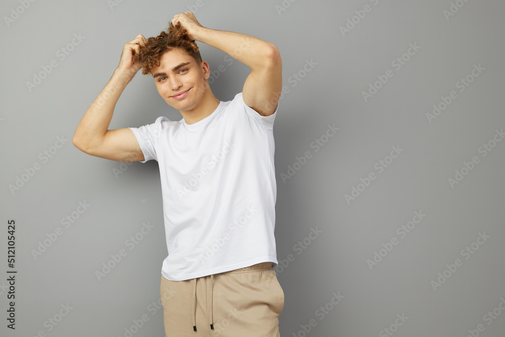 an attractive young man stands on a gray background in a white cotton T-shirt and looks at the camera with his hands on his curly hair an attractive young man stands on a gray background in a white cotton T-shirt and looks at the camera with his hands on his curly hair