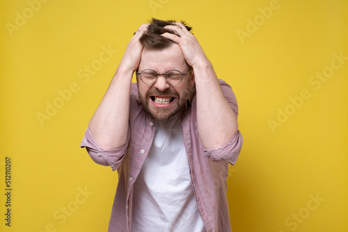 Young man is irritated, he is tearing the hair on his head in stress and showing teeth. Concept of mental health. Yellow background.
