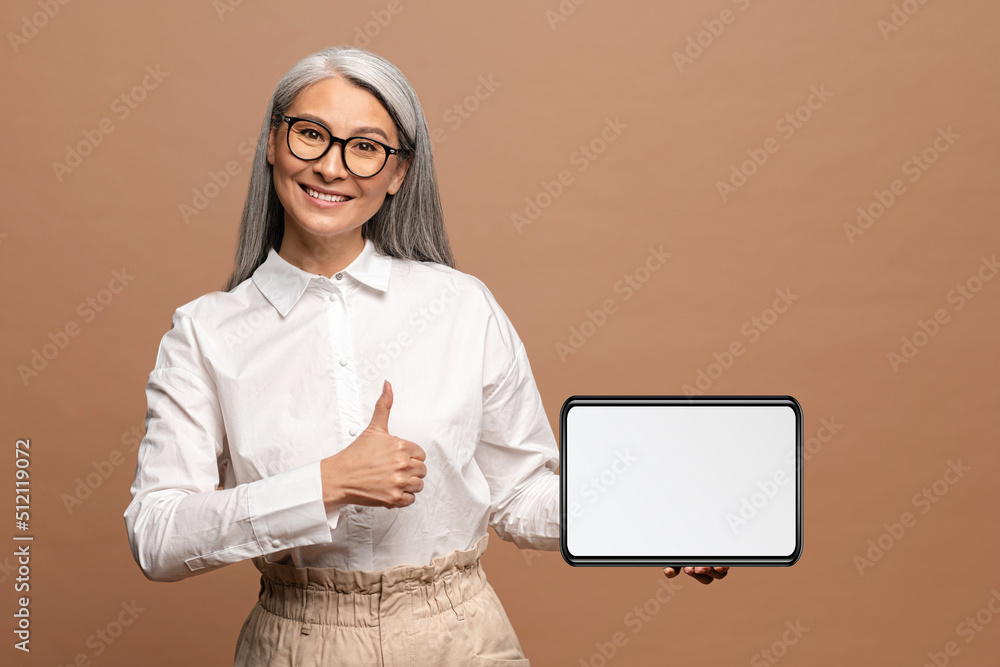 Portrait of the confident senior woman holding tablet and looking at the camera with big finger gesture. Indoor studio shot isolated on beige background