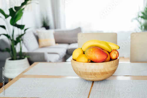 Kitchen counter table with focus on bamboo bowl with exotic fruits on it with blurred background of modern cozy living room with couch and green plants. Open space home interior design. Copy space
