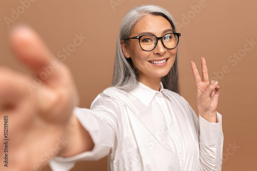 Lady smiling and looks at the camera isolated on beige. Cheerful senior asian woman taking selfie  beautiful mature female holding hand in victory gesture