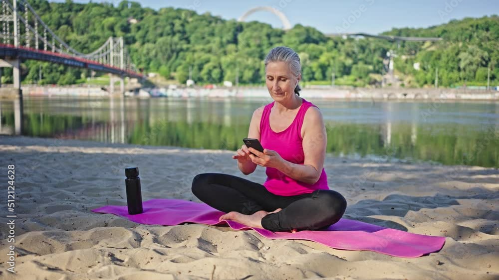 Senior woman sits on pink mat looking in smartphone by river Senior woman sits on pink mat looking in smartphone by river