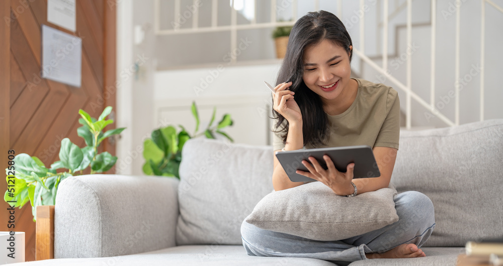 Beautiful attractive young Asian woman sitting on gray sofa and using digital tablet at living room in the morning. Beautiful attractive young Asian woman sitting on gray sofa and using digital tablet at living room in the morning.