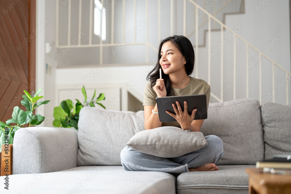 Beautiful attractive young Asian woman sitting on gray sofa and using digital tablet at living room in the morning. Beautiful attractive young Asian woman sitting on gray sofa and using digital tablet at living room in the morning.