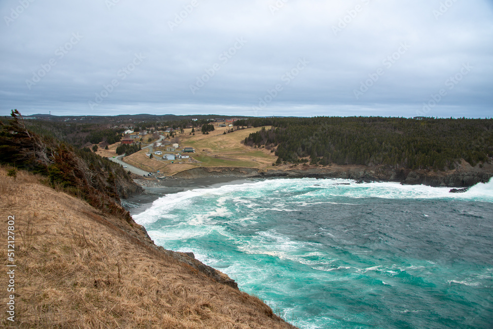 An aerial view of Middle Cove Beach with waves rolling onshore. There are small farmhouses with large grassy meadows, forest, and green grass. There's a gravel road leading to the beach and cove.