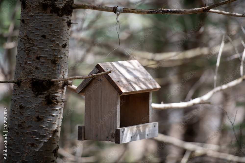A weathered wooden bird feeder hanging in an evergreen tree in a wooded area. The birdhouse has an open side with seeds on the tray.  The handmade shelter is for wild birds to feed on in winter. 