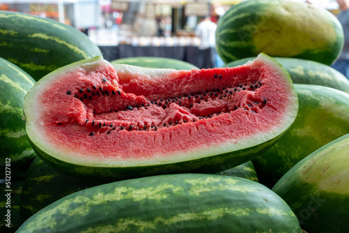 Fresh elongated watermelon with a transverse cut showing a pink chunky inside with lots of black seeds and striped green and yellow exterior rind with lots of whole melons on a table for sale.   