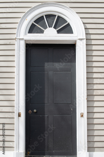The exterior of a vintage building with beige colored narrow clapboard cape cod siding. There's a black wooden door with a thick white decorative trim. A half circle transom window hangs over the door
