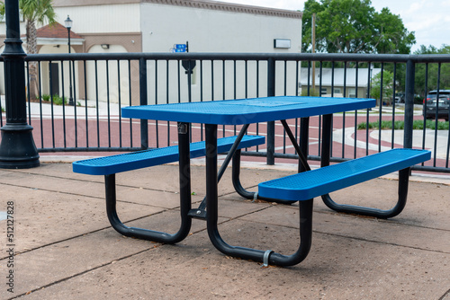 A vibrant blue and black empty metal picnic table on a ceramic patio of a beer garden. There's a white concrete shop in the background with trees and a parking area. 