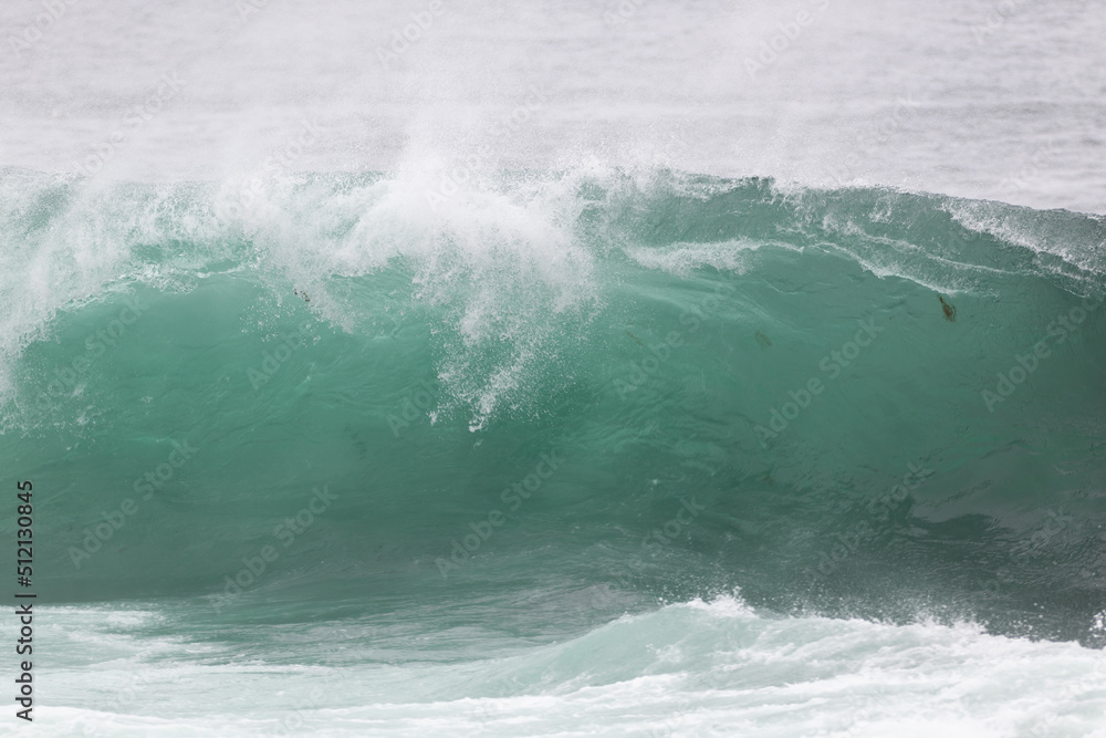 An angry teal green color massive rip curl of a wave as it barrels rolls along the ocean. The white mist and froth from the wave are foamy and fluffy. The ocean spray is coming off the top of the wave