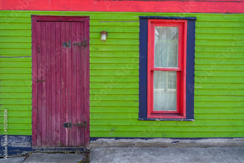 A wooden purple storm door with big black hinges on a lime green exterior wall of a building with red and blue trim around the two windows. The colorful window is a vintage double hung closed glass.