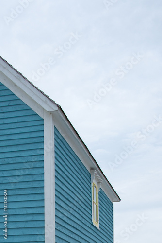 An upward view of a vintage blue wooden building with thick white trim, a small double hung window, with four panes of glass, and a frame shingled roof. The roof has white trim and black shingles. © Dolores  Harvey
