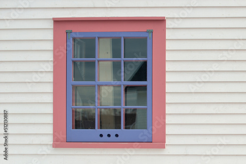 A single vintage storm window with 12 small panes  a purple wooden frame  and pink trim in a white wooden clapboard building. The exterior wall is made of narrow pine horizontal clapboard siding.