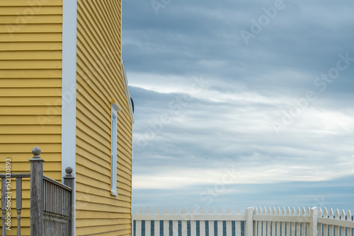 A yellow country style wooden building with a white picket fence attached and enclosing the boundary of a yard overlooking the blue ocean with a cloudy sky. The house has a wood cape cod siding. 