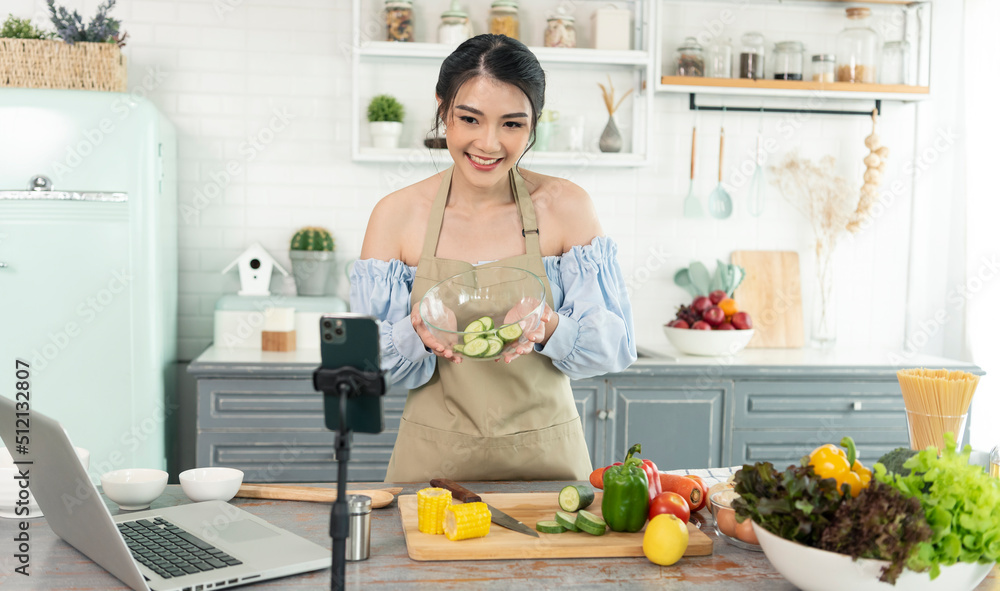 Asian woman food blogger cooking salad in front of smartphone camera while recording vlog video and live streaming at home in kitchen.
