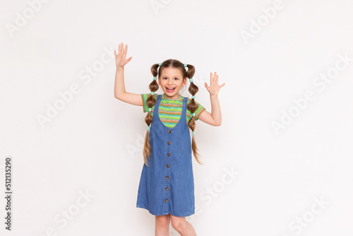 A little girl with a funny hairstyle points with her palms at your advertisement on a white isolated background. A child in a summer denim sundress.