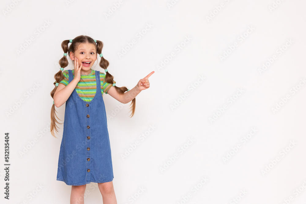 The child points sideways with his index finger at your advertisement on a white isolated background. A beautiful girl in a denim sundress. Copy Space The child points sideways with his index finger at your advertisement on a white isolated background. A beautiful girl in a denim sundress. Copy Space